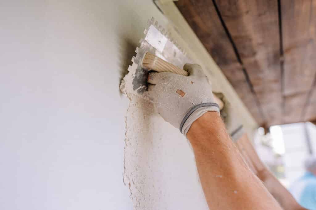 Main gantée de travail appliquant de l'enduit sur un mur blanc avec une truelle crantée, sous un plafond en bois.