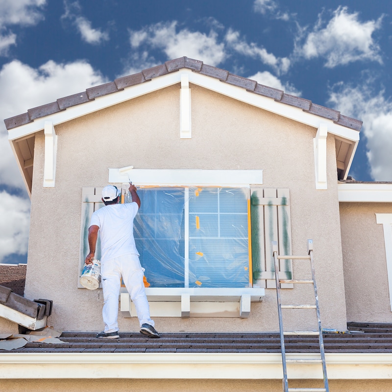 Peintre professionnel en tenue blanche appliquant une couche de peinture sur la façade d'une maison beige, au-dessus d'une fenêtre protégée.