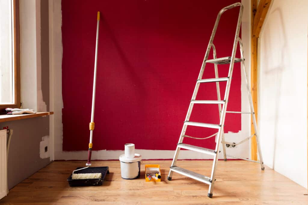 Pièce en rénovation avec mur rouge vif fraîchement peint, échelle, rouleaux et pots de peinture blanche au sol en bois.
