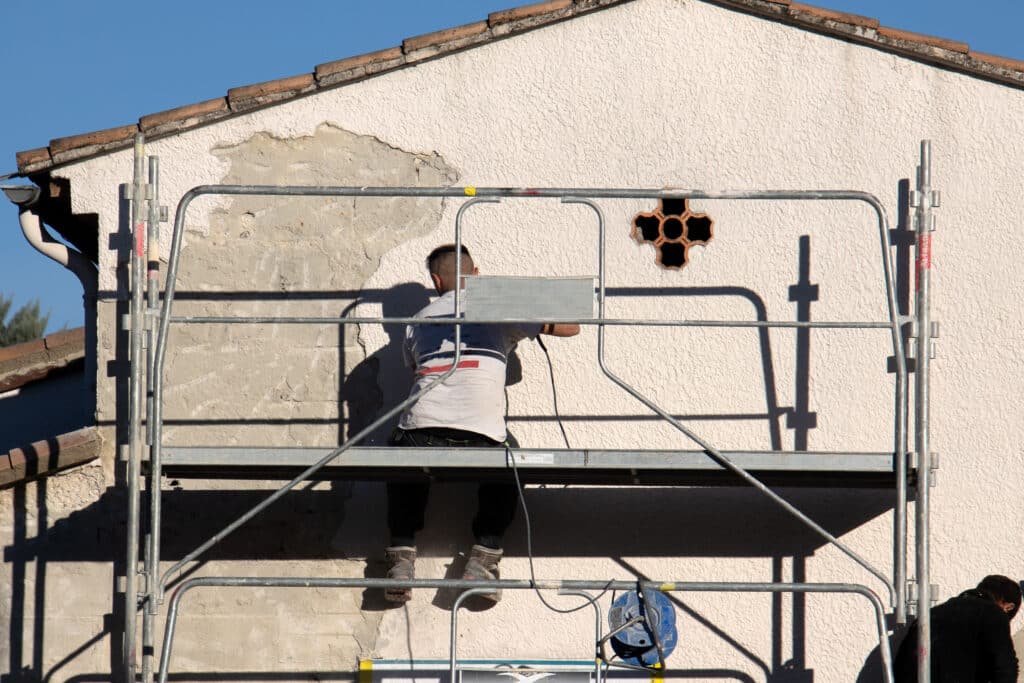 Ouvrier sur échafaudage rénove un mur extérieur crépi d'une maison. Le vieux plâtre s'écaille. Un vent hexagonal est visible. Matériel ALTRAS.